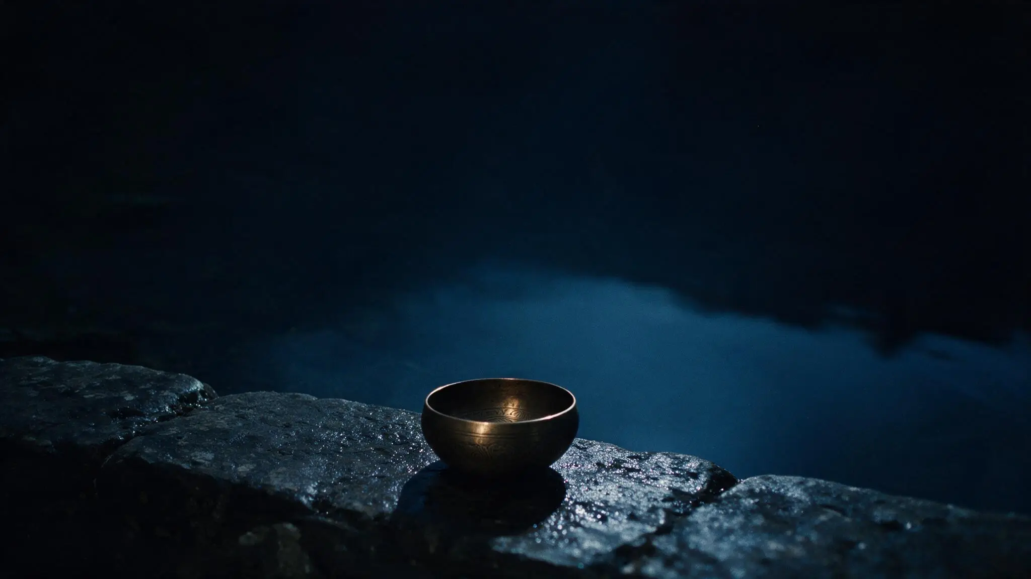A wet dark stone edge at the water, with one bronze bowl set to the side and still water beyond.