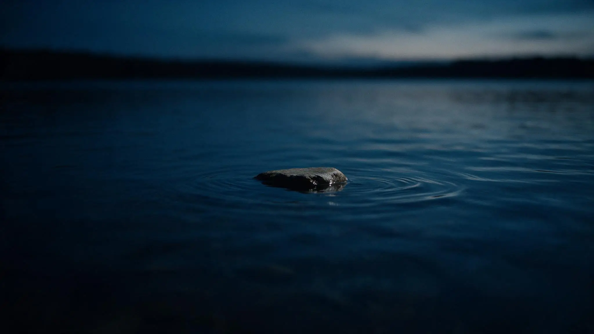 A single low flat stone standing just above the water halfway across, with faint rings around it.
