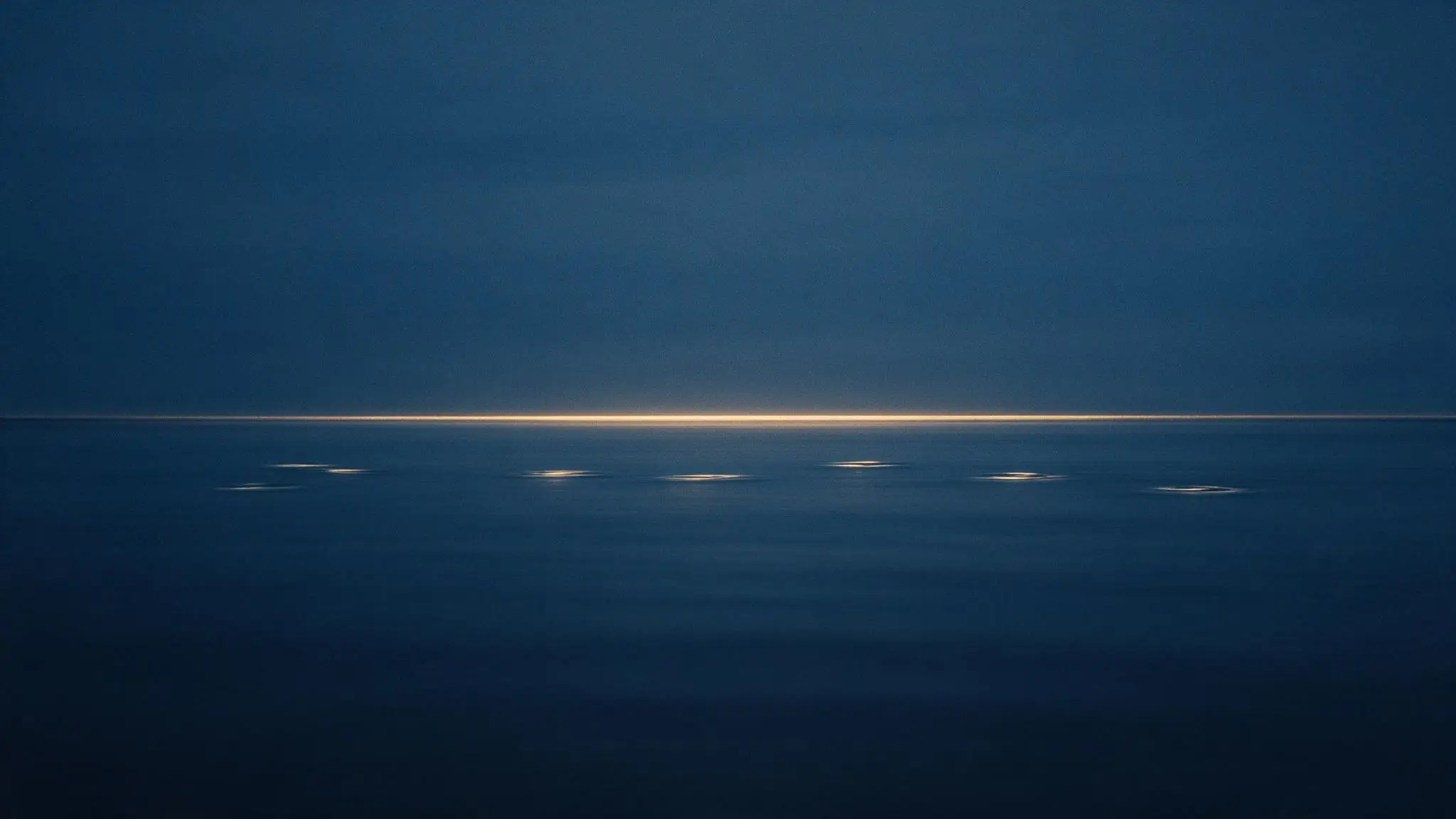 A calm sea at blue hour with one thin band of gold light on the horizon.